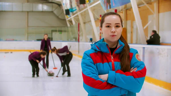 Curlingträning inomhus - domaren i blå jacka står på ishallen och tittar in i kameran - hennes elever spelar curling på bakgrunden — Stockfoto