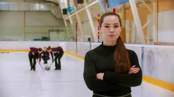 Curling utbildning - domaren som står på ishallen tittar i kameran med korsade händer - hennes elever spelar curling på bakgrunden — Stockfoto