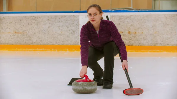 Curling entrenamiento en pista de hielo - una joven con gafas de pie en la pista de patinaje sosteniendo una piedra y cepillo —  Fotos de Stock