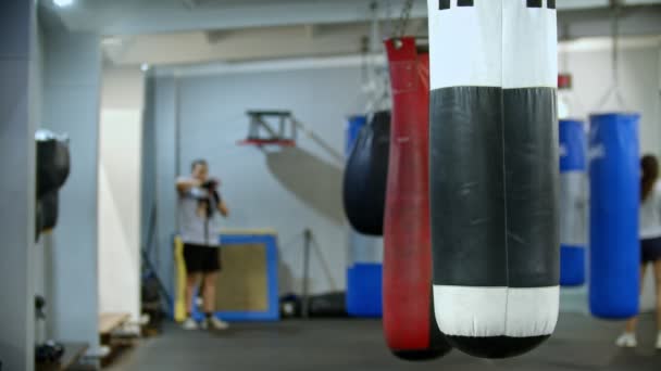 Egy bokszzsák lóg oldalt a tornateremben - az ember bekötözi a kezét egy kötéssel — Stock videók
