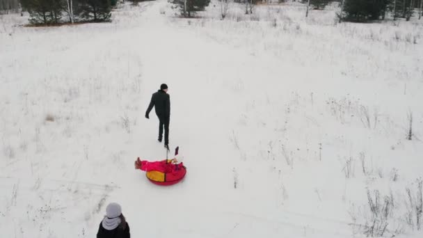 Щаслива сім'я грає на зимовому полі батько їде на надувних санях, а його дружина і дочка бігають за ними — стокове відео