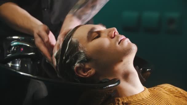 Ein junger Mann lässt sich im Friseursalon die Haare mit Shampoo waschen — Stockvideo