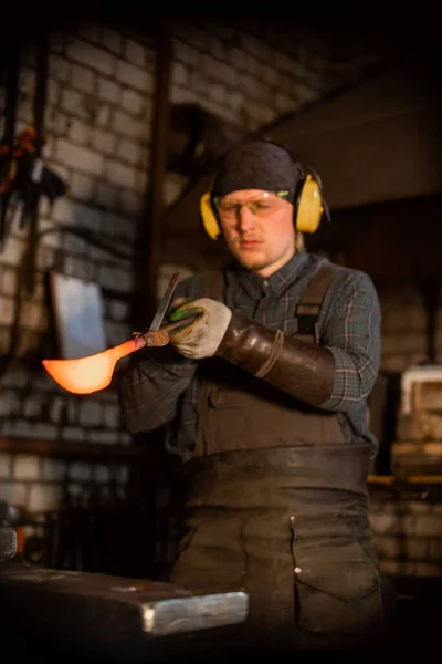 Un uomo nella protezione delle cuffie che tiene il campione di coltello in metallo caldo nel forcipe — Foto Stock