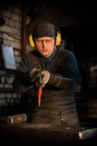 Un uomo nella protezione di cuffie e occhiali forgiando un coltello - tenendo il campione in pinza — Foto Stock