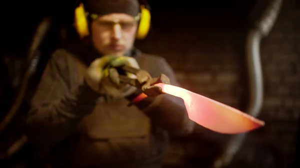 Um ferreiro homem fazendo uma faca com um martelo à procura de imperfeições — Fotografia de Stock