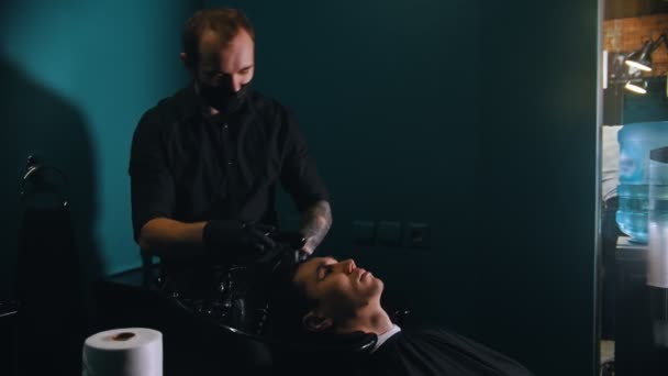 Man barber in black mask with tattoes washing his client hair — Stock Video