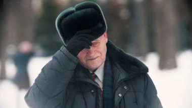 An old smiling man takes off his hat and cleaning it up from the snow