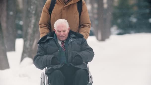 Молодой человек гуляет на улице с дедушкой в инвалидном кресле в зимнее время — стоковое видео