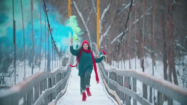 Dos jóvenes mujeres felices corriendo en el puente nevado sosteniendo coloridas bombas de humo — Vídeo de stock