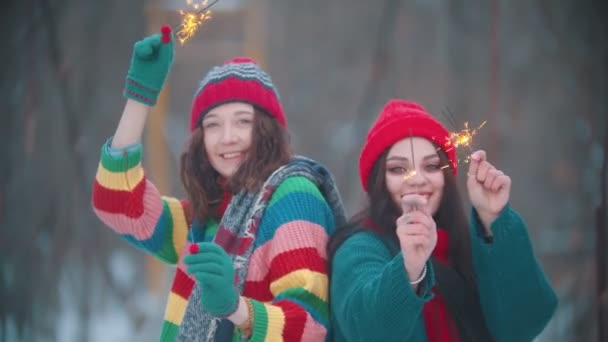 Duas jovens mulheres sorridentes brincando no inverno com brilhos iluminados e olhando para a câmera — Vídeo de Stock