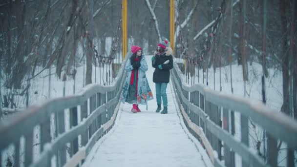 Дві молоді жінки ходять і розмовляють по засніженому мосту - п'ють гарячі напої з термоса — стокове відео