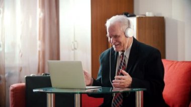 Elderly grandfather - old grandfather in headphones smiling and looking at laptop screen in his rooom