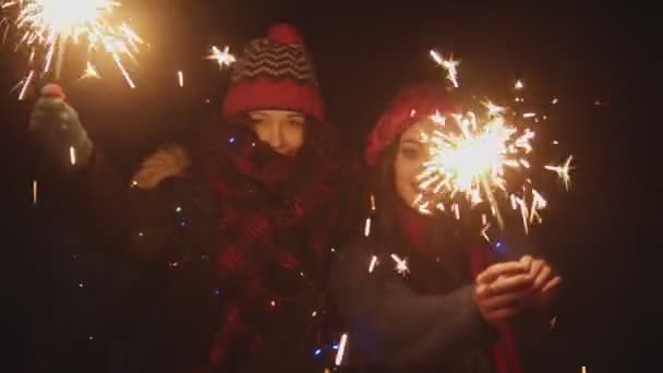 Zwei junge hübsche Frauen, die nachts draußen mit Wunderkerzen spielen und in die Kamera schauen — Stockvideo