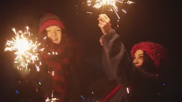 Duas jovens mulheres sorridentes amigos brincando com faíscas à noite e olhando para a câmera — Vídeo de Stock