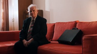 Elderly grandfather - grandfather is sitting on a sofa with a suitcase and touching a hand over his face