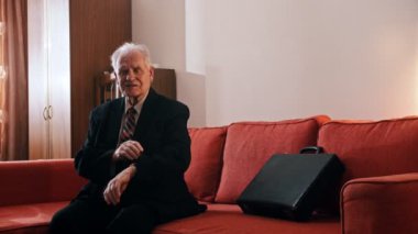 Elderly grandfather - grandfather is sitting on the couch with a suitcase and checking the time on the swatch in his room