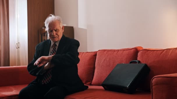 Літній дід - дід сидить на дивані з валізою і перевіряє час на годиннику — стокове відео