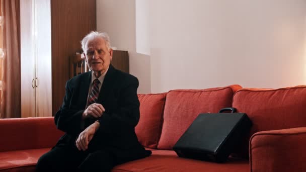 Anciano abuelo - abuelo está sentado en el sofá con una maleta y comprobar la hora en la muestra en su habitación — Vídeo de stock