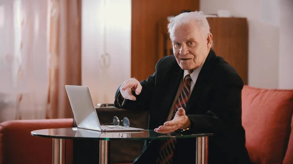 An old businessman in glasses pointing at the laptop and looking in the camera — Stockfoto