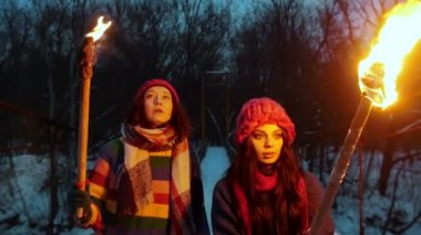 İki genç bayan arkadaş, ellerinde meşalelerle köprüde yürüyorlar ve etrafa bakınıyorlar.