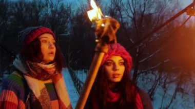 Akşamleyin elinde meşalelerle köprüde yürüyen iki genç bayan arkadaş meşaleleri tutuyorlar.