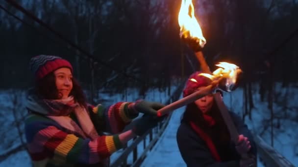 Két fiatal, mosolygó nő harcol tüzes fáklyákkal a téli erdőben. — Stock videók