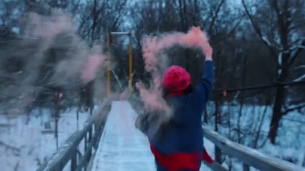 Eine junge Frau läuft auf der schneebedeckten Brücke mit einer rosa Rauchbombe — Stockvideo