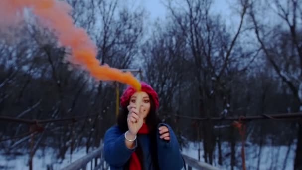 Una joven hermosa mujer caminando sobre el puente nevado sosteniendo una bomba de humo naranja — Vídeo de stock