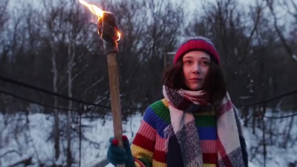 Una joven caminando sobre el puente nevado en el bosque sosteniendo una antorcha encendida — Vídeos de Stock