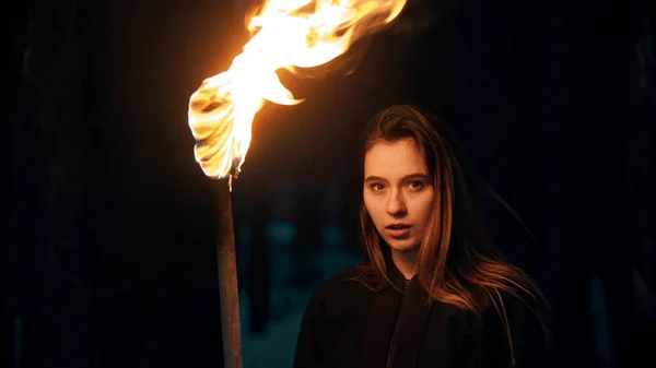 Молодая красивая женщина с огненным факелом в ночном лесу смотрит в камеру — стоковое фото