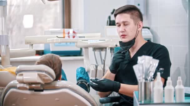 Молодой стоматолог надевает маску для лица перед тем, как проверить рот мальчика — стоковое видео