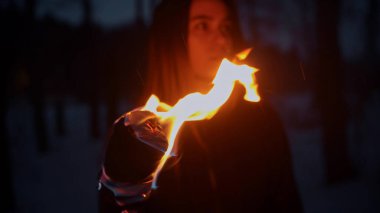 Genç bir kadın, elinde bir ateş meşalesiyle geceleri dışarıda dikiliyor ve yan tarafa bakıyor.