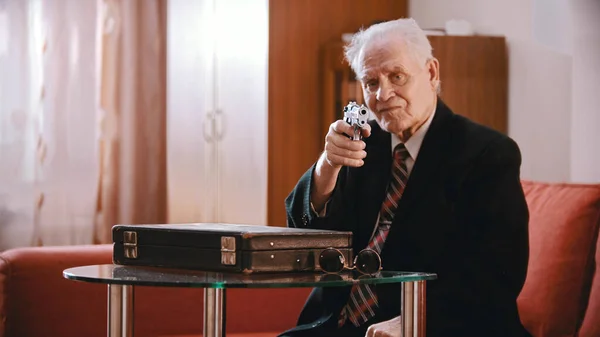Egy öreg üzletember ül a bőrönd mellett, és pisztollyal céloz a kamerába. — Stock Fotó
