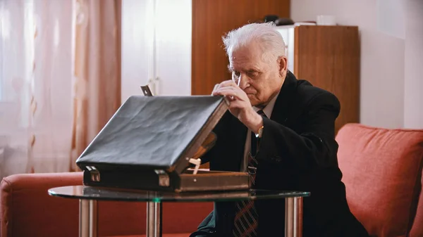 An old businessman sitting on the sofa and opening a suitcase — Stock Photo, Image