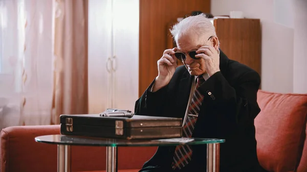 Starý obchodník sedící u kufru a nasazující si brýle — Stock fotografie