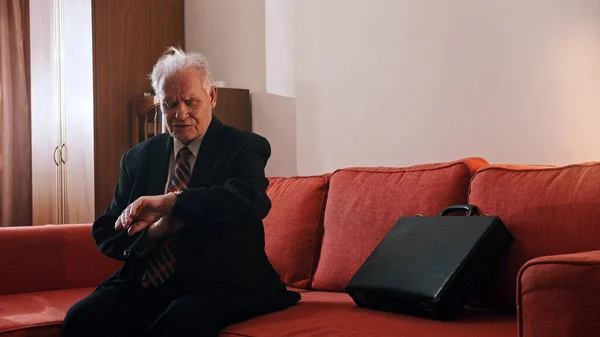 Ein alter Mann Großvater sitzt auf dem Sofa und schaut auf seine Handuhr — Stockfoto