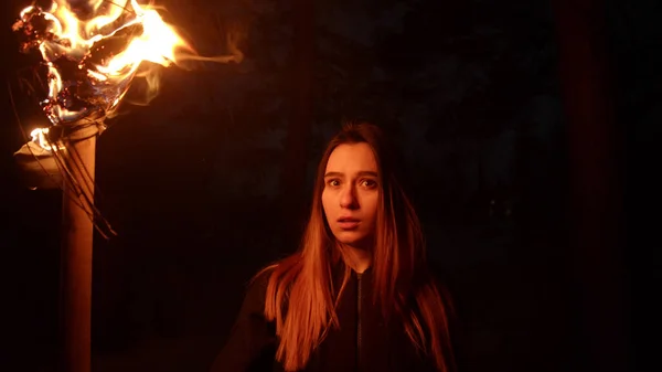 Young woman holding fire torch standing in dark forest and looking at something with fear