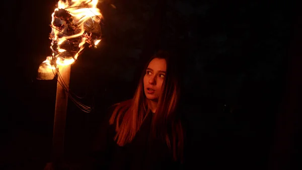 Jovem mulher segurando fogo tocha em pé na floresta escura à noite e olhando para algo com medo — Fotografia de Stock