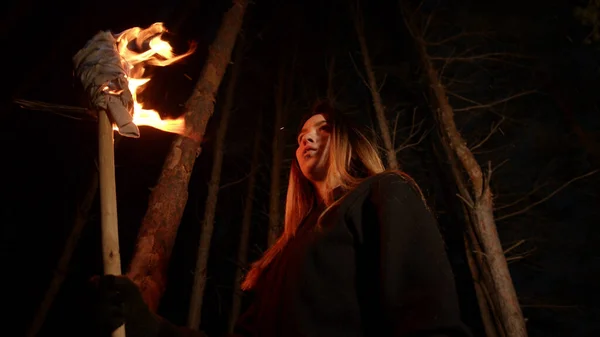 Молодая потерянная женщина с длинными волосами, стоящая ночью в лесу с факелом ручной работы — стоковое фото