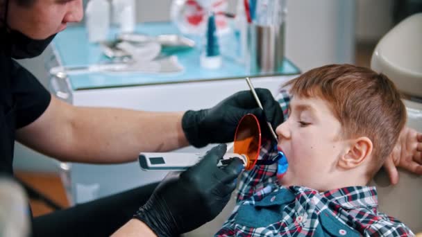 Маленький мальчик с вырезанными зубами - кладет фотополимерную лампу в рот и включает ее — стоковое видео