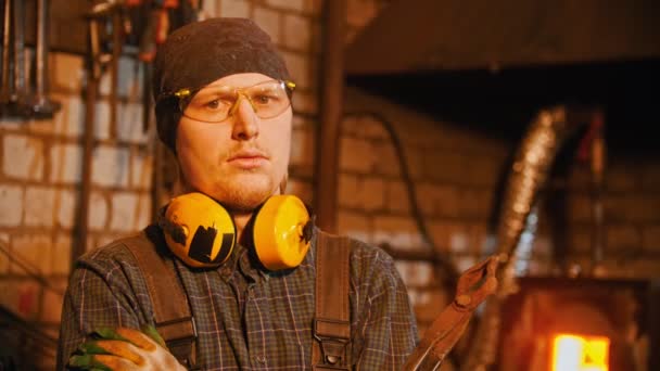 Indústria de forjamento - um ferreiro homem em óculos e fones de ouvido segurando fórceps e olhando para a câmera — Vídeo de Stock