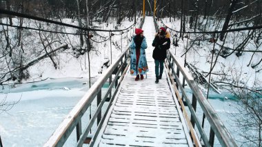 Two young women students drinking hot drinks and walk on the snowy bridge in the winter forest