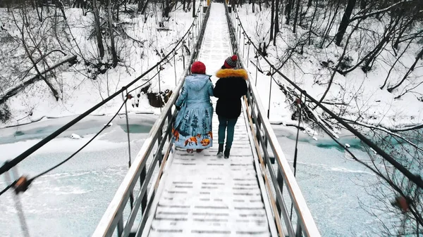 Две молодые женщины в пуховиках пьют горячие напитки и ходят по снежному мосту — стоковое фото
