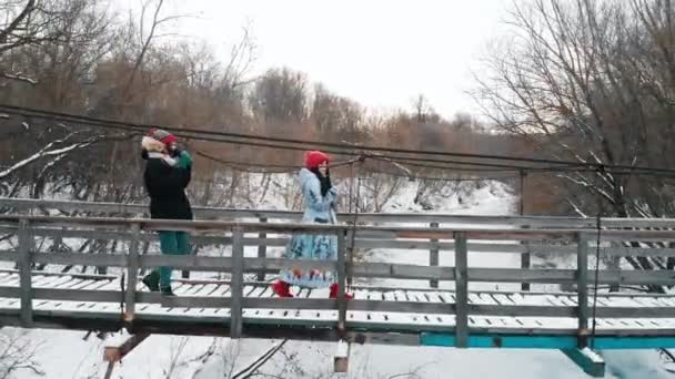 Duas jovens mulheres bebem bebidas quentes da garrafa térmica e caminham na ponte — Vídeo de Stock