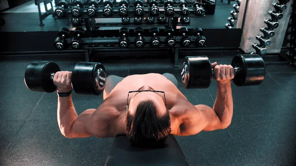 Un hombre culturista en gafas bombeando los músculos de sus manos tumbado en el soporte en el gimnasio — Foto de Stock