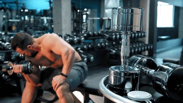 A man bodybuilder pumping his hand muscles sitting in the sports gym - dumbbells on the foreground — Stock Photo, Image