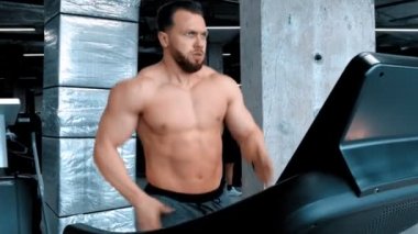 Sakallı, çekici, genç bir vücut geliştirici modern spor salonunda bir çabayla koşu pistinde koşuyor.