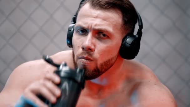 En attraktiv man boxare i hörlurar sitter på bänken och dricksvatten - tittar i kameran — Stockvideo