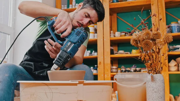 Керамика - мастер в зеленой футболке сушит глиняную чашу со строительным феном — стоковое фото