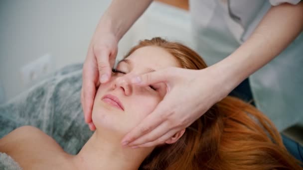 Massaggio - massaggiatrice massaggiatrice che massaggia il viso di una donna dai capelli rossi — Video Stock